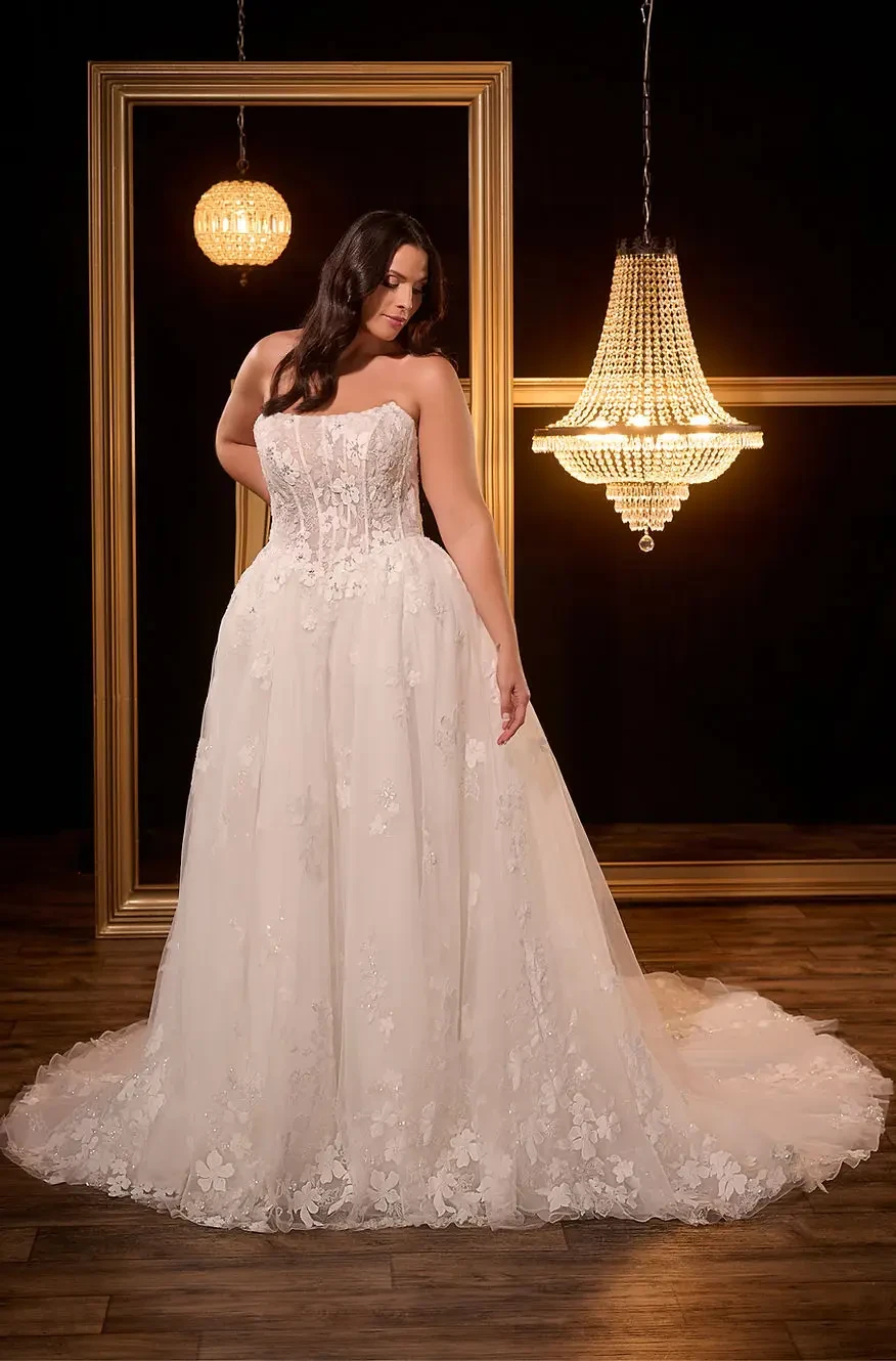 A woman in a strapless, lace wedding dress stands on a wooden floor. She is framed by golden frames and chandeliers, with a serene expression.
