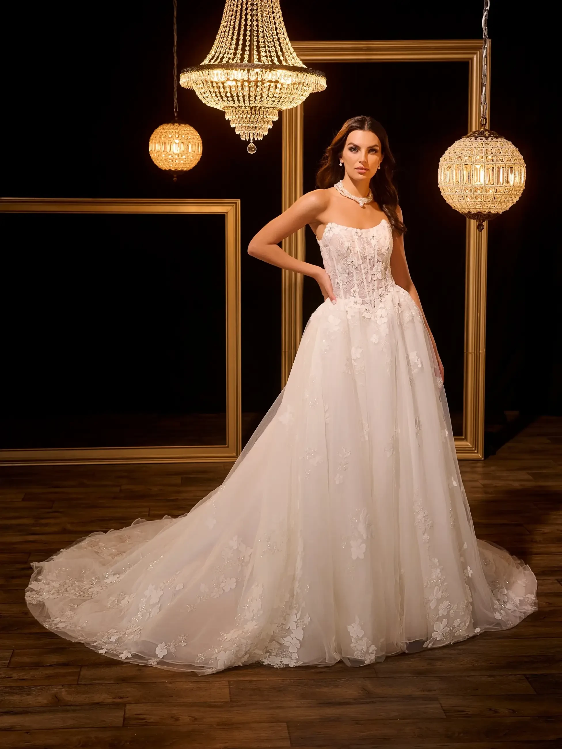 A woman stands confidently in an elegant white lace wedding gown with a long train. She's framed by chandeliers and gold picture frames on a dark backdrop.