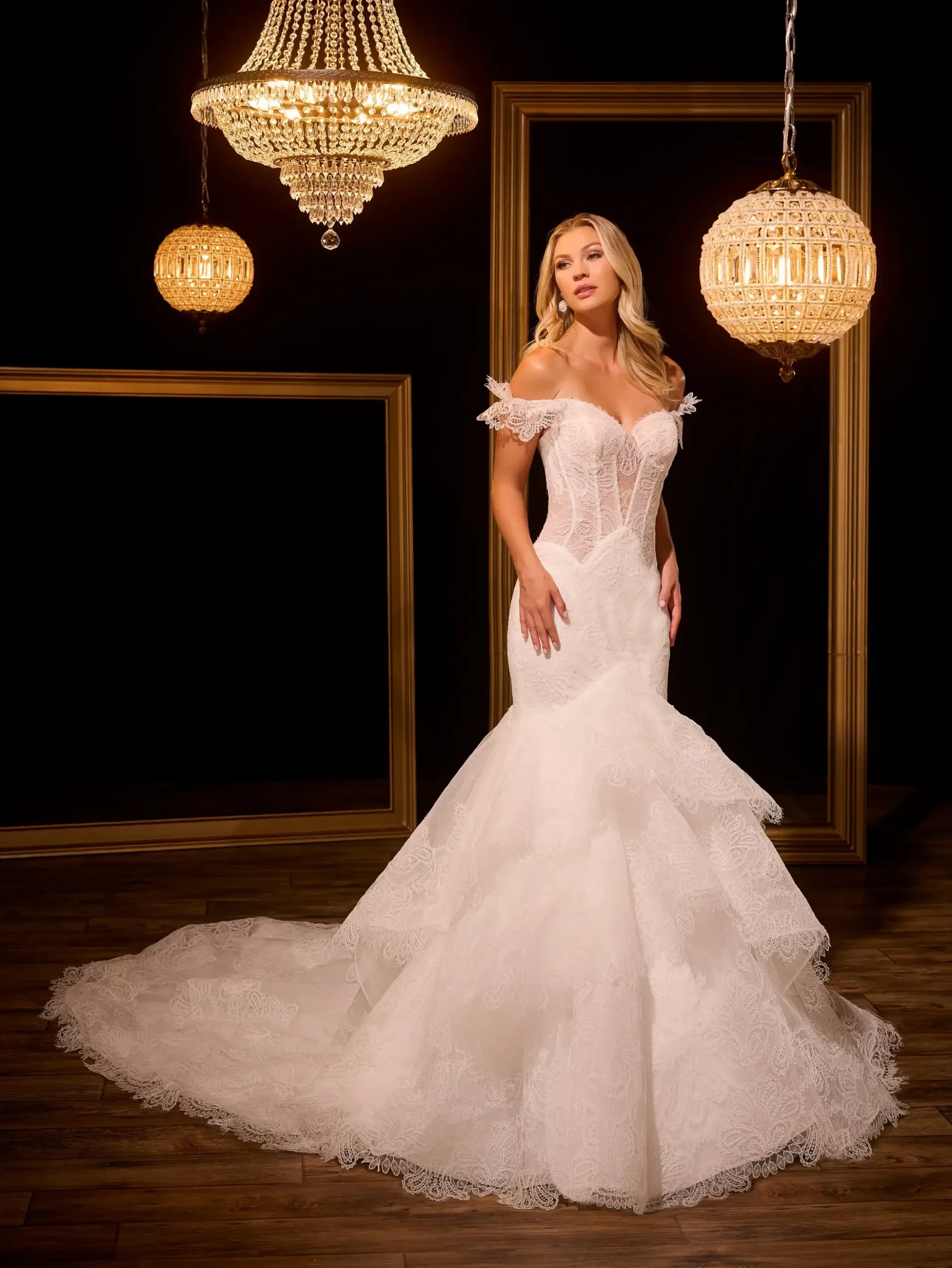 A woman in an elegant, off-shoulder lace wedding gown with a mermaid silhouette stands on a wooden floor. She is surrounded by ornate chandeliers, creating a warm, luxurious ambiance.