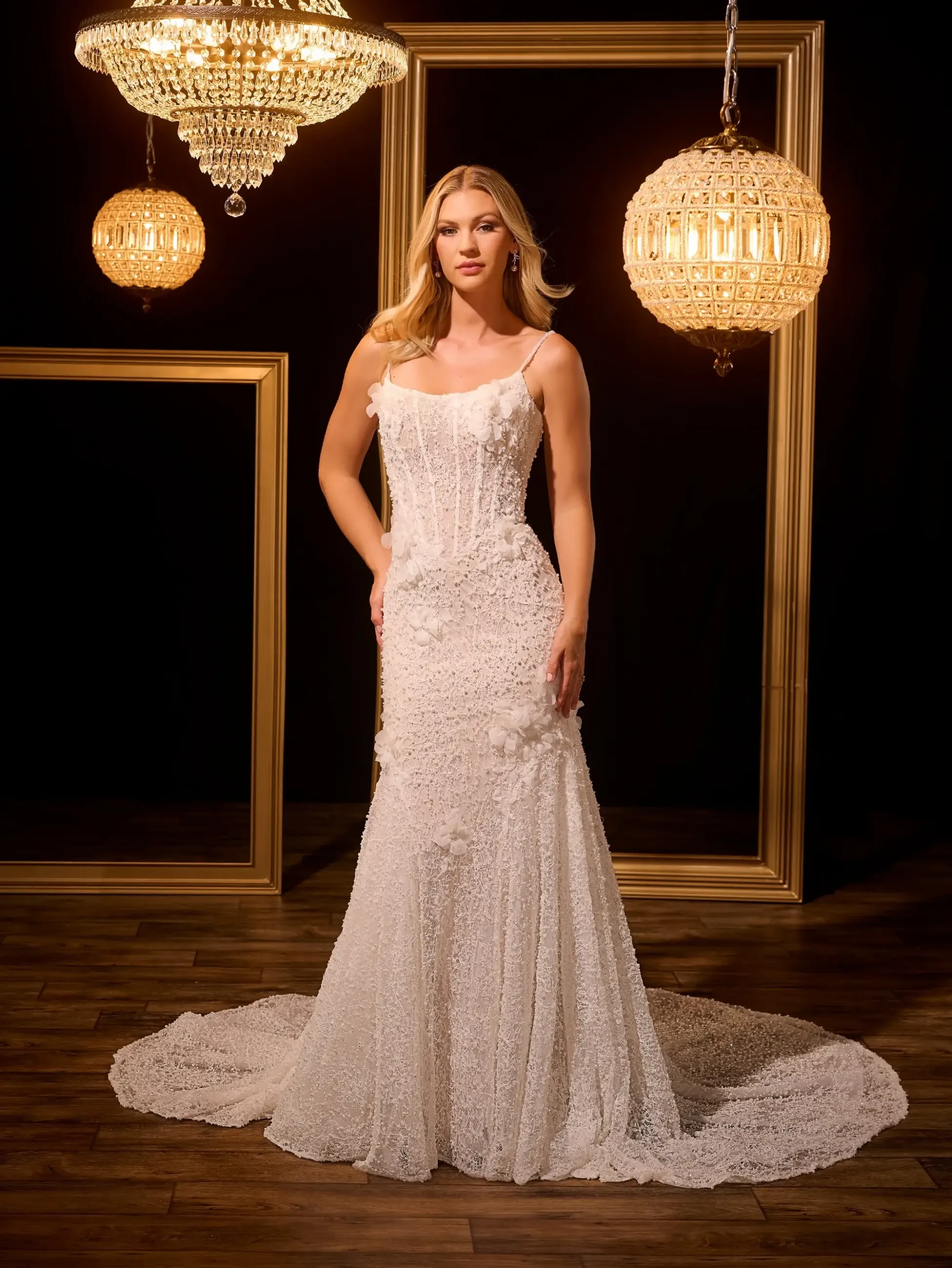A woman in an elegant, beaded white dress stands confidently between ornate chandeliers and gold frames, on a wooden floor, exuding sophistication.