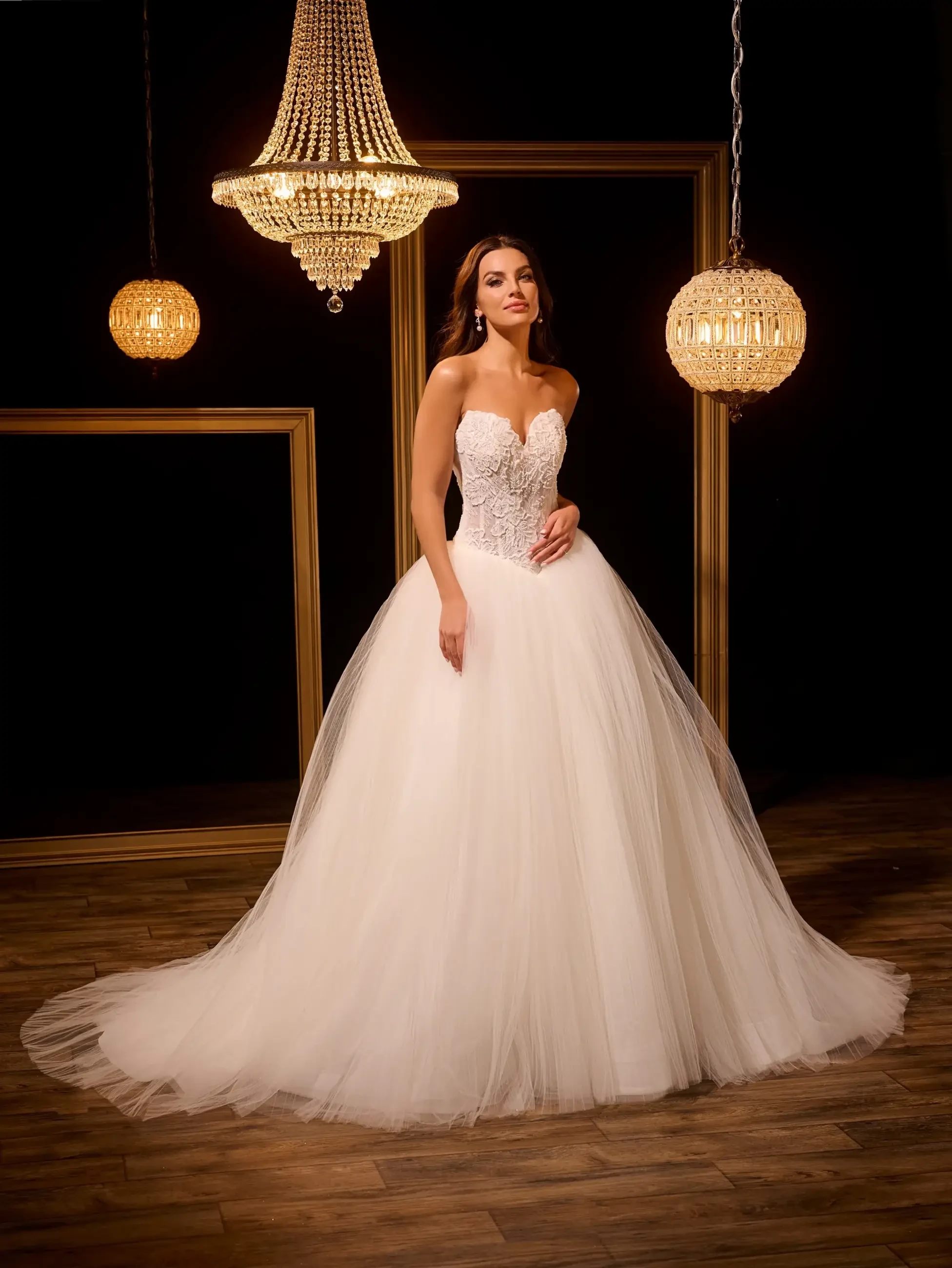 A bride in a strapless white wedding gown with a fitted bodice and a full tulle skirt, standing in an elegantly lit room with chandeliers.