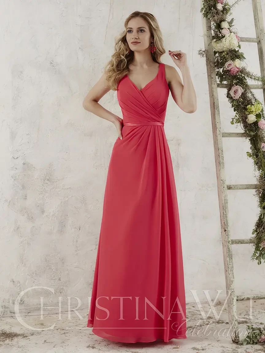 A woman in a coral-colored maxi dress with a V-neckline and spaghetti straps, standing elegantly against a light background with floral decorations.
