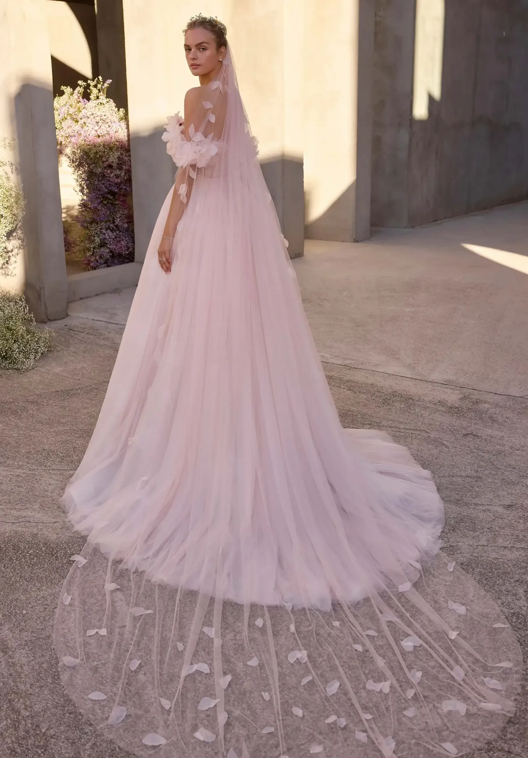 Model is wearing a romantic A-line wedding gown with layers of gathered tulle and organza, a plunging neckline, 3D floral off-the-shoulder sleeves, and a satin bow at the waist.