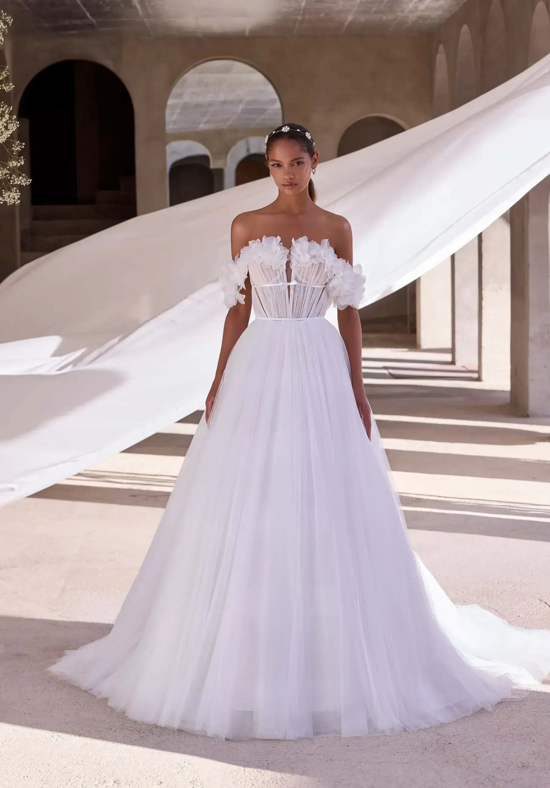 Model is wearing a romantic A-line wedding gown with layers of gathered tulle and organza, a plunging neckline, 3D floral off-the-shoulder sleeves, and a satin bow at the waist.