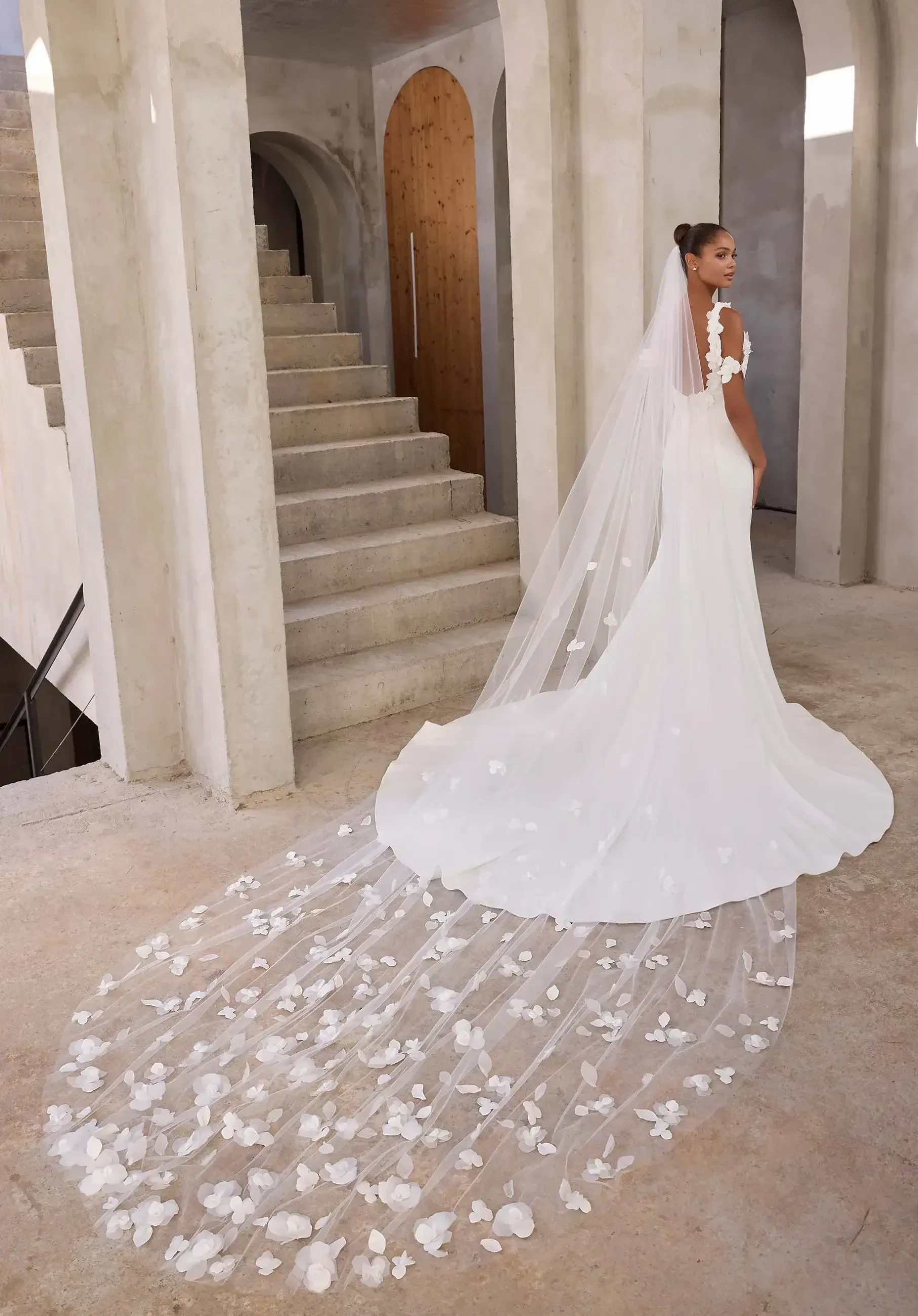 Model is wearing a sleek crepe fit-and-flare gown with floral appliqués, a detachable off-the-shoulder drape, scoop back, thigh-high slit, and sweeping train.