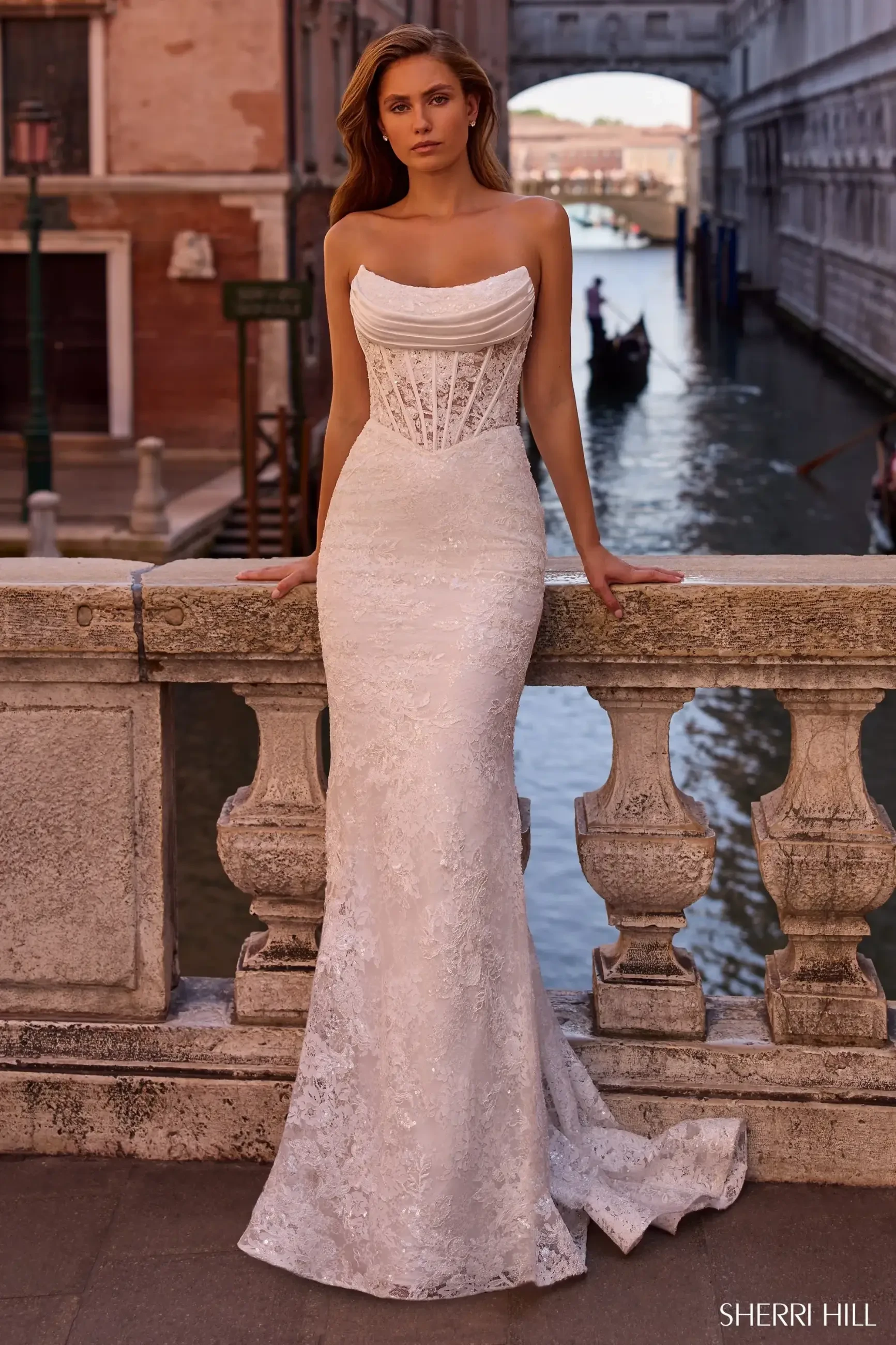 A model in a stunning white dress with a fitted bodice and flowing skirt, posing on a stone railing overlooking a canal in Venice, Italy, with gondolas in the background.