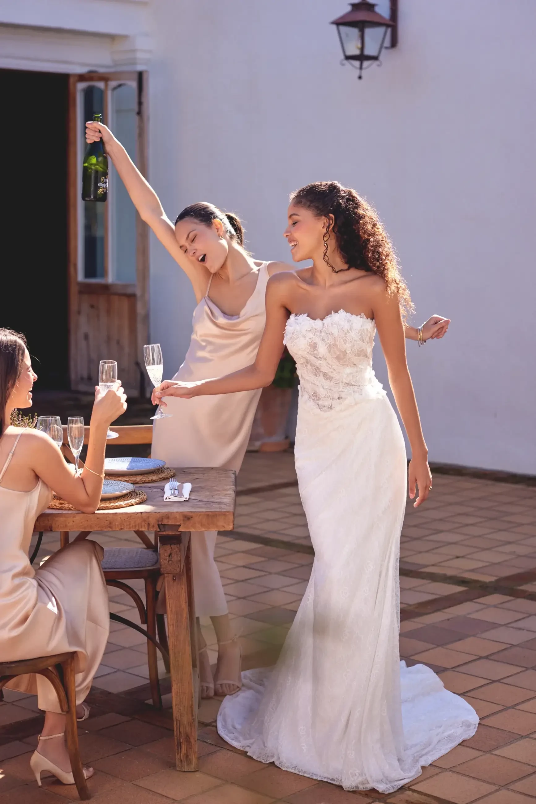 Bride in white lace gown smiles beside two women in champagne dresses. One woman raises a champagne bottle; the other sits holding a glass, celebrating outdoors.