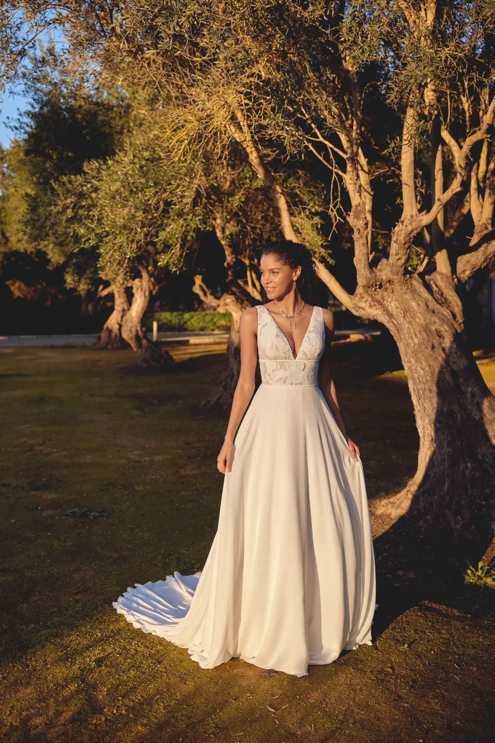 Bride in elegant flowing gown stands amidst sunlit trees, exuding tranquility and happiness. The scene captures a warm, golden ambiance.