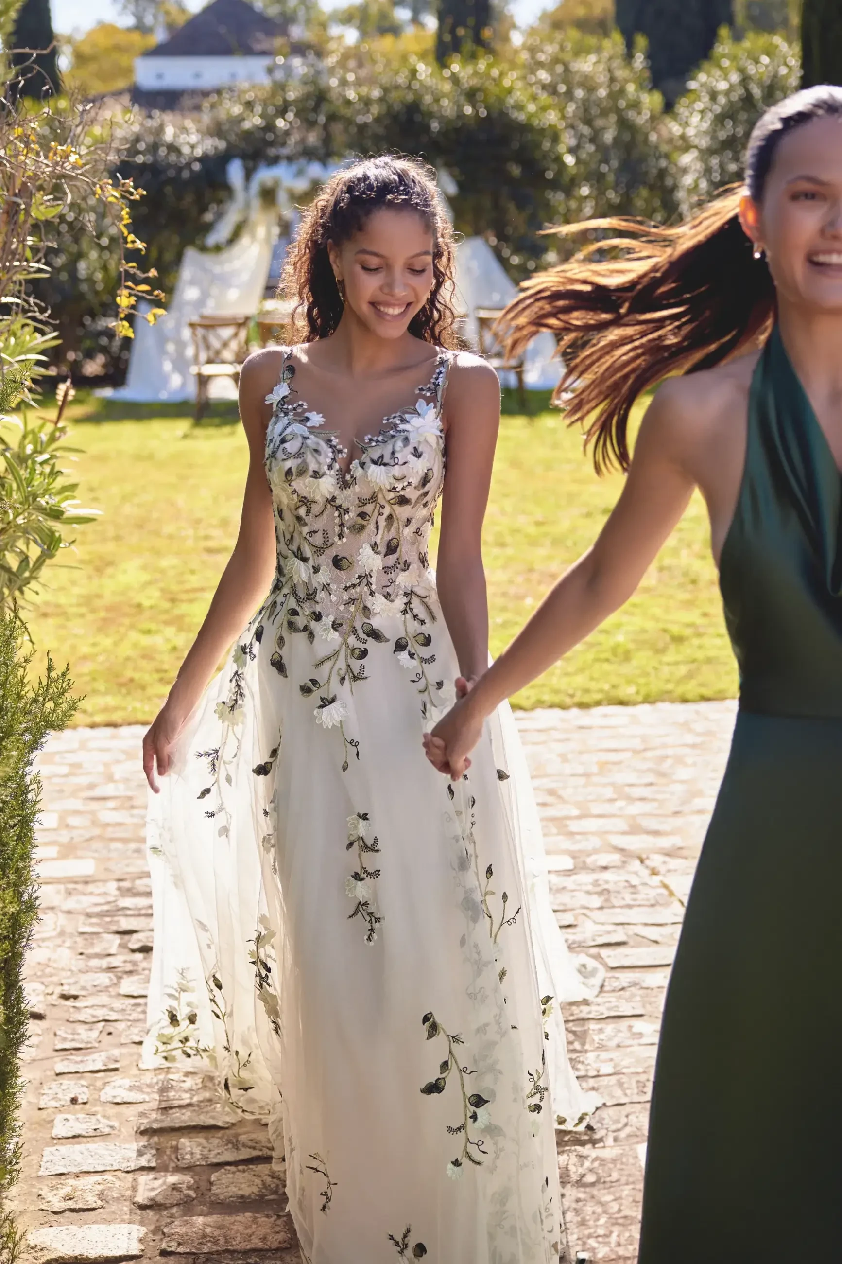 A smiling woman in a floral embellished wedding dress walks hand-in-hand with another woman in a green dress outdoors.