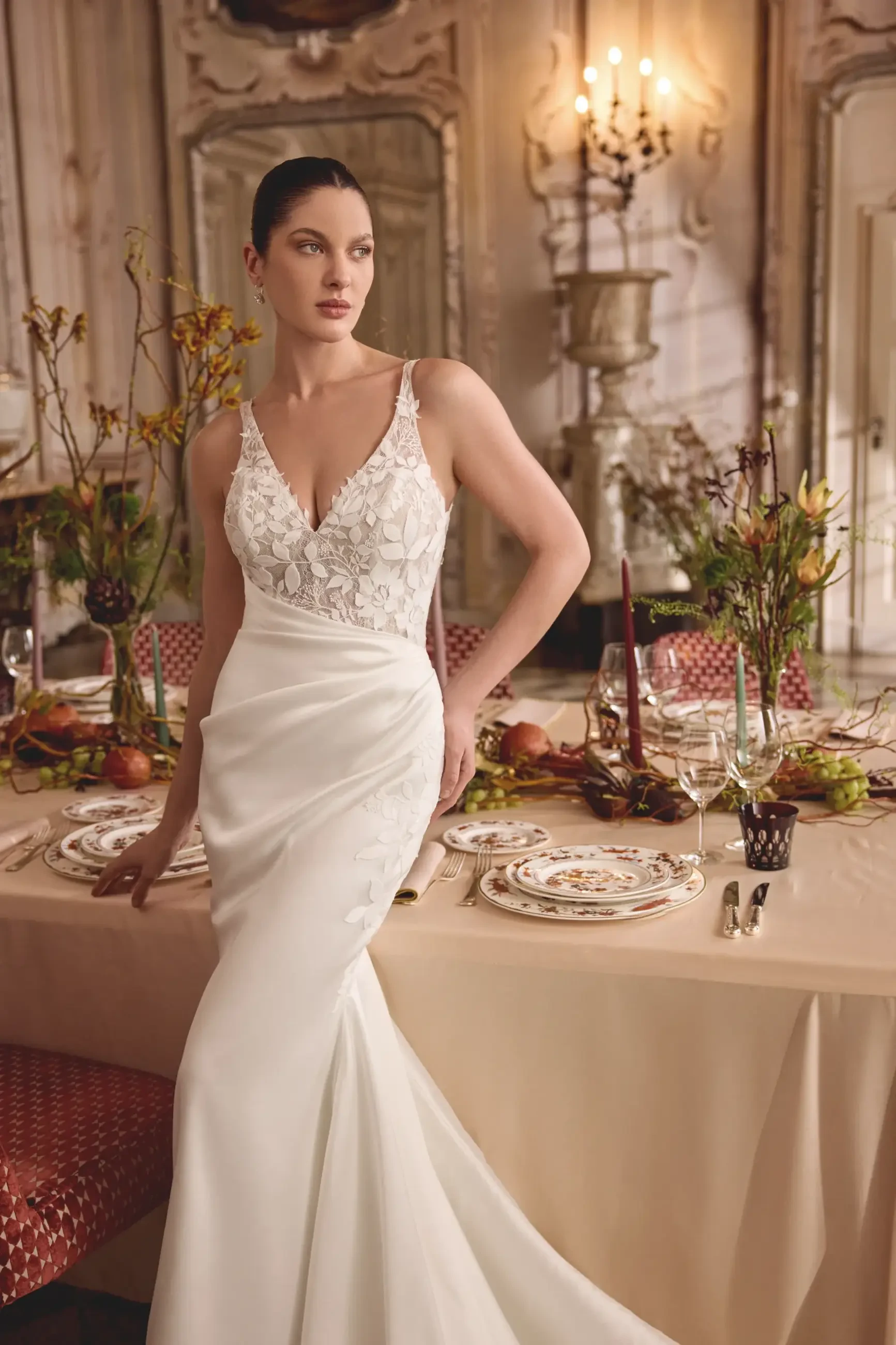 A woman in an elegant white gown with lace detailing is posing next to a beautifully set dining table decorated with autumn-themed elements. The background features ornate decor and soft lighting.
