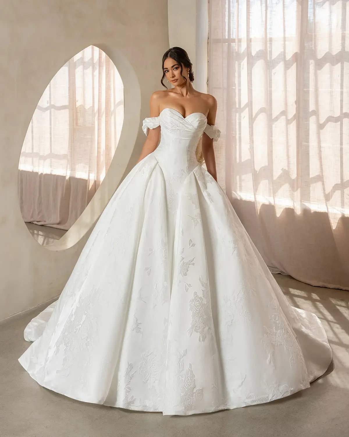 Bride in an elegant off-shoulder white gown with intricate details stands in sunlit room, exuding grace. Soft shadows and neutral tones enhance the scene.
