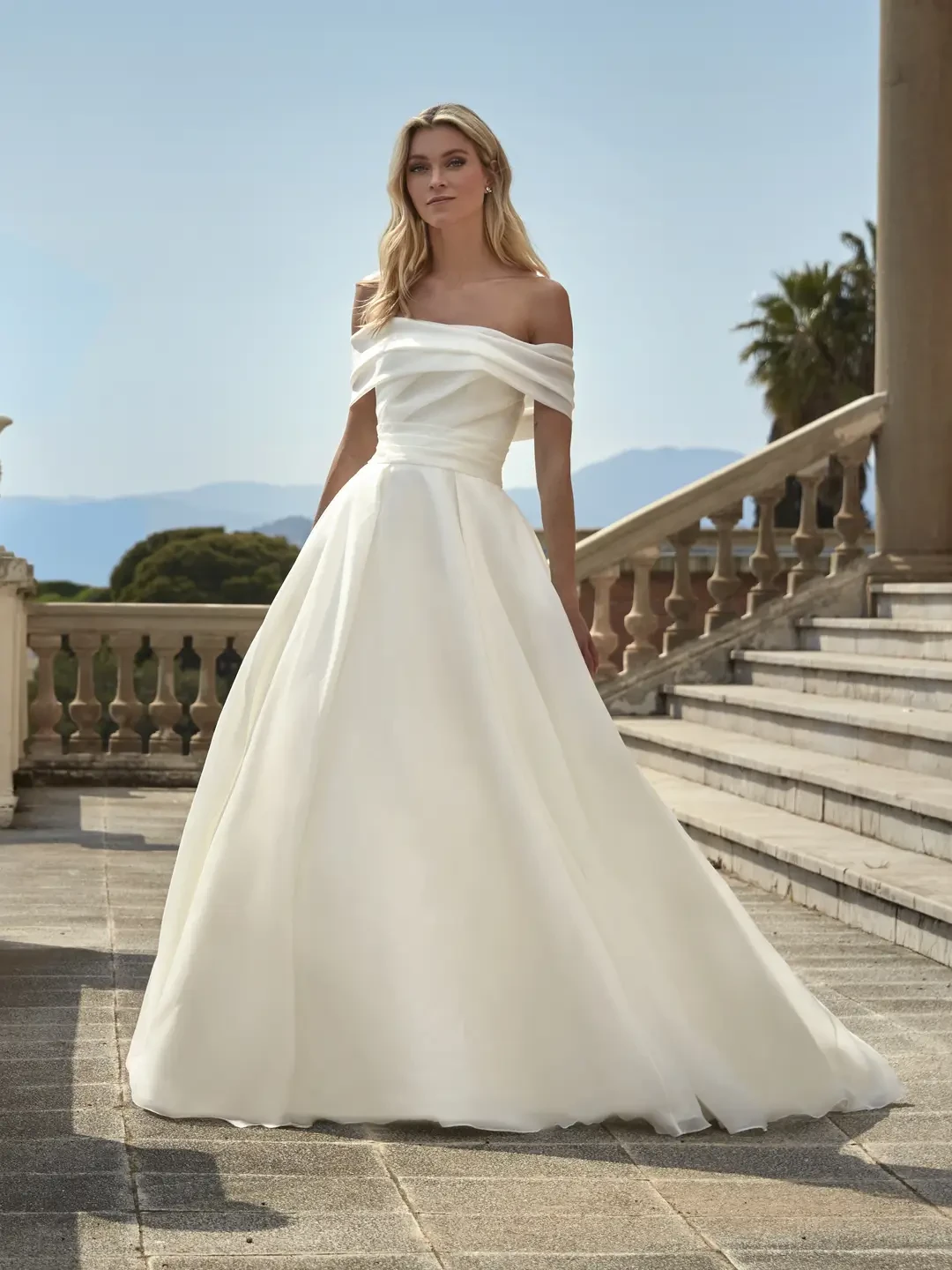 A woman stands elegantly outdoors, wearing an off-the-shoulder white wedding gown. Background features stone stairs, railings, and a clear blue sky.